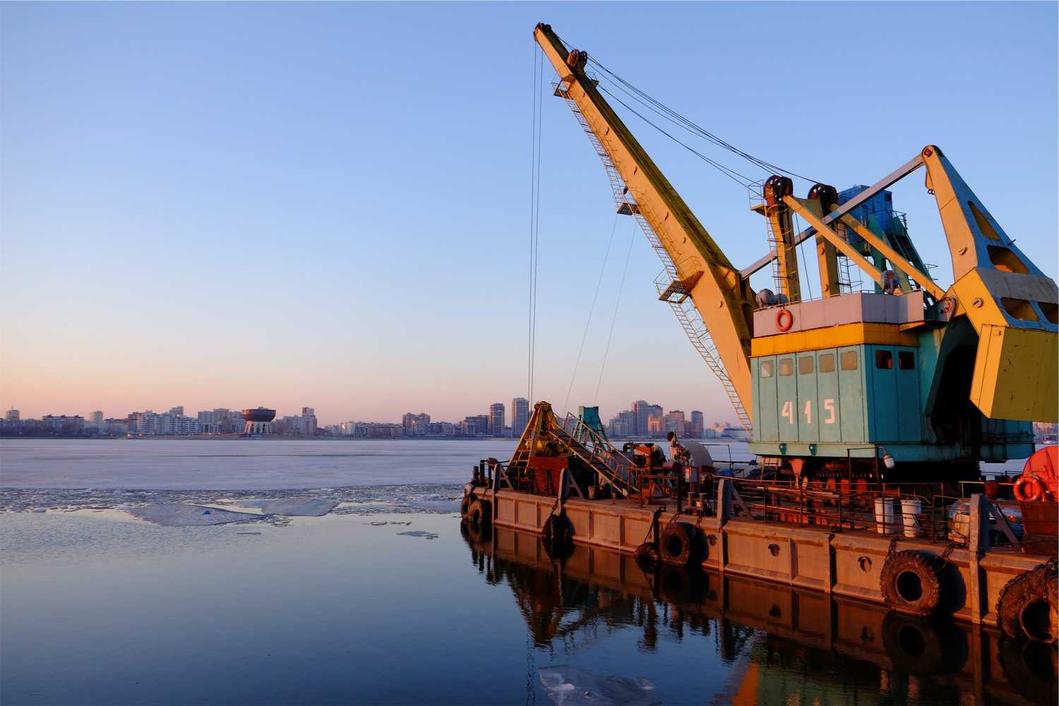 A massive yellow and blue crane sits on a barge parked on a frozen riv…